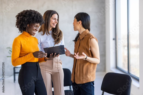 A diverse trio of professional women engage in a dynamic conversation in a bright office, reviewing a document on a clipboard.