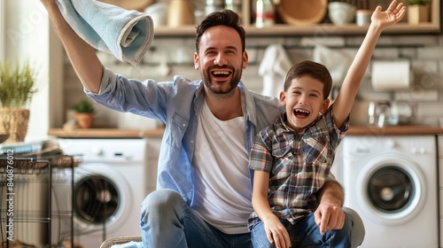 A happy father and son smile and throw towels in the air while sitting on the floor near the washing machine in their kitchen at home.