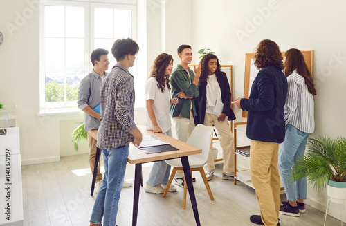 Young male teacher standing with college or high school students, talking and discussing something near a blackboard in the classroom during a lesson. Education, learning and teaching concept.