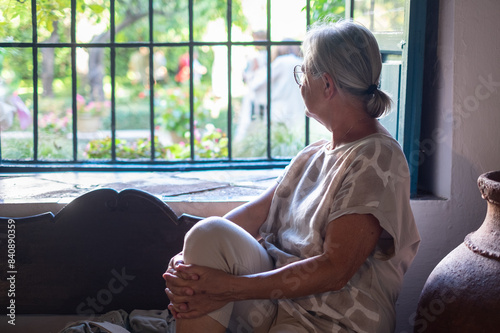 Mature senior woman sitting indoors at the window looking out at the green garden