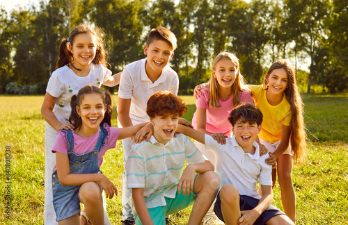 Portrait of a happy smiling children friends sitting on the green grass together in the park outdoors and looking cheerful at the camera hugging and having fun. Kids summer camp and holidays concept.