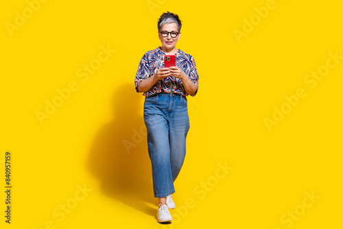 Full length photo of lovely senior lady walk hold device dressed stylish colorful garment isolated on yellow color background