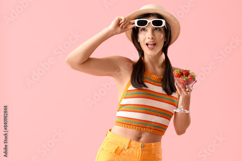 Surprised young woman holding glass with fresh strawberries on pink background