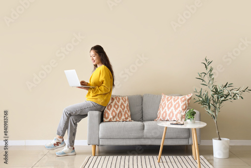 Young Asian woman using laptop on grey sofa near beige wall