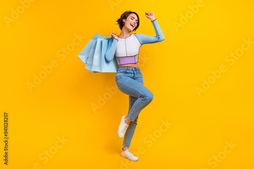 Full body photo of delighted cheerful lady hold mall shop bags raise fist success isolated on yellow color background