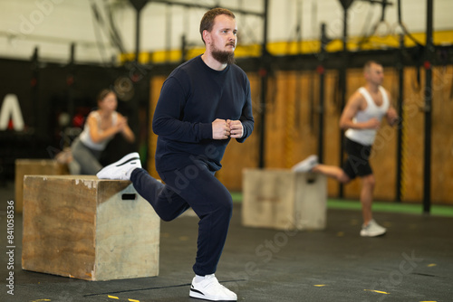 Concentrated young bearded man in sport wear performing Bulgarian split squats on wooden plyometric box during intense bodyweight training at gym..