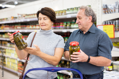 Elderly married couple choosing canned cucumbers together in the grocery section of supermarket