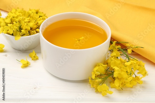 Rapeseed oil in bowl and beautiful yellow flowers on white wooden table