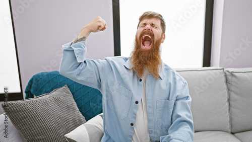 Cheerful young redhead man confidently flexing strong biceps, radiating joy with a cool smile, while comfortably sitting in his homely living room.