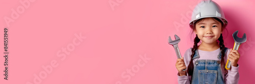 A cute dark-haired Asian girl holds construction tools in her hands on a pink isolated background. Banner, place for text