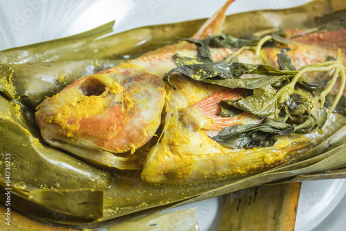 Indonesian food: pepes ikan nila (steamed nile tilapia with spices). Spices are smeared on tilapia's body, wrapped with banana leave, and then steamed. On top of a glass plate and white tablecloth.