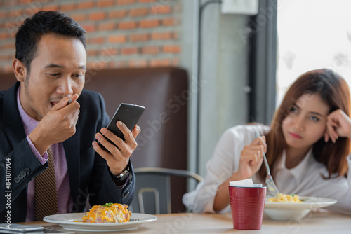 A man is looking at his phone while a woman is sitting across from him looking bored.