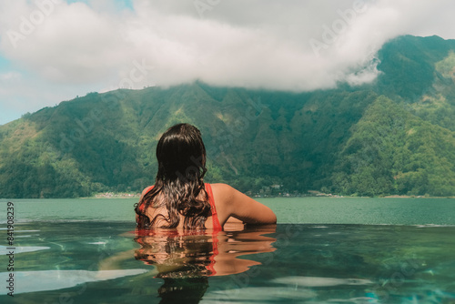 A young woman on vacation relaxing in the summer at hot thermal springs in a mountainous area. She sits in the pool and looks into the distance at the hills and lake that is located in front of her.