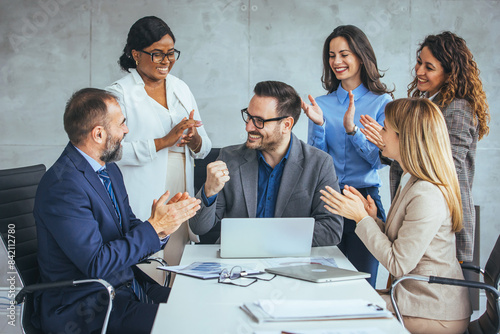 A group of diverse business professionals celebrate success, clapping for a smiling male colleague seated with a laptop in a modern office.