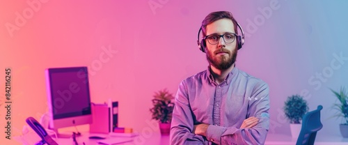 Confident male software engineer wearing headphones and glasses standing in front of a computer in an office and looking at the camera with crossed arms.