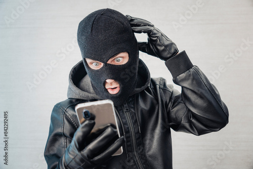 Young adult in black clothes with hidden face. Ill-intended fraudster uses mobile. Fraudster calls. Scam. Mobile racket. Hacker hijacks by phone. Cellphone account fraud.