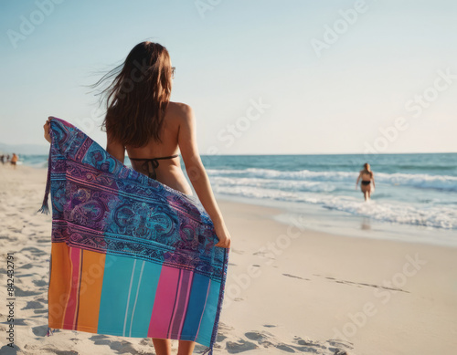 Un telo da mare con un design astratto, ricco di colori accesi e forme fluide, è posato su una spiaggia tranquilla, con il mare calmo che si estende all'orizzonte. 