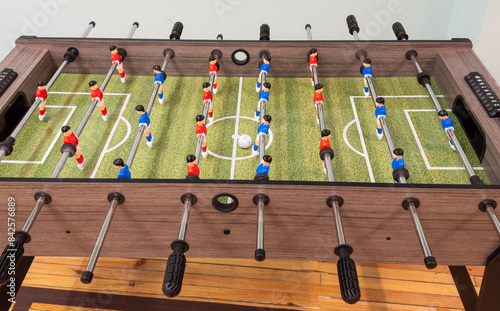 Classic Foosball Table Game With Players in Red and Blue Jerseys During Daytime Indoor Match. Close-up view of foosball table featuring red and blue jersey players positioned for daytime indoor game.