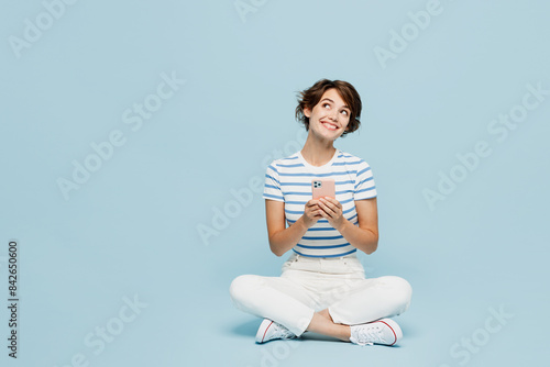 Full body young minded smiling woman wears striped t-shirt casual clothes sitting hold in hand use mobile cell phone isolated on plain pastel light blue background studio portrait. Lifestyle concept.