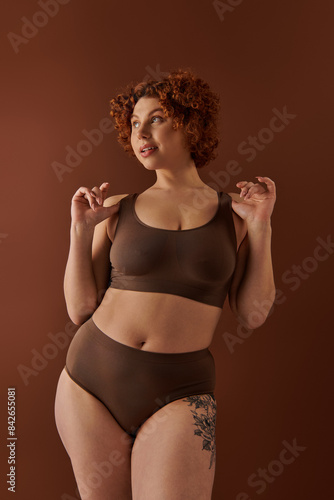A young and curvy redhead woman in a brown bikini poses gracefully on a warm-toned brown background.