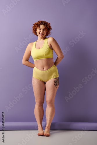 Curvy redhead woman flaunting a yellow bikini in front of a vibrant purple wall.