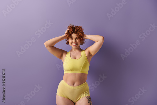 A young, curvy redhead woman confidently poses in a yellow bikini against a striking purple background.