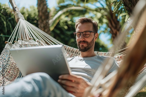 Young hipster man holding digital tablet computer and working remotely from a hammock outdoors. Vacation, holiday, workation, remotely working, online learning, freelance. Digital nomad lifestyle