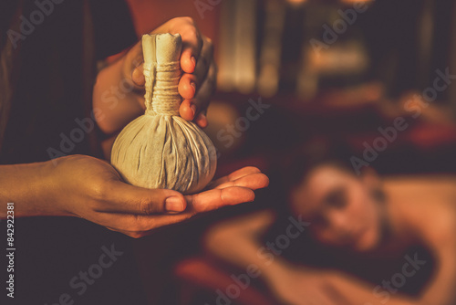 Hot herbal ball spa massage body treatment with masseur's hand hold or show herb bag at spa. Tranquil and serenity of aromatherapy recreation in warm lighting of candles at spa salon. Quiescent