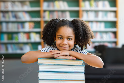 Smiling, young African American girl, studying in the library, resting her chin on her hands atop a stack of books. Early education and learning concept.