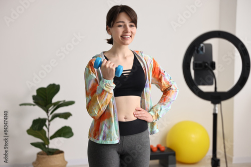 Happy sports blogger holding dumbbell while streaming online fitness lesson with smartphone at home