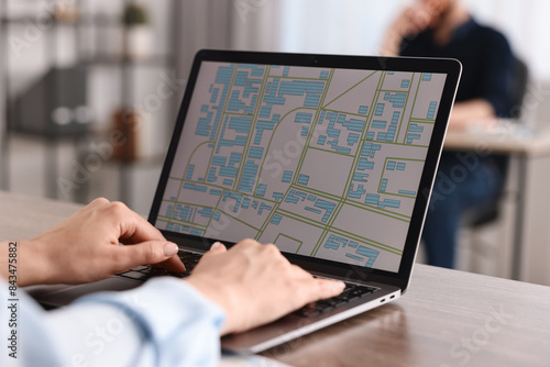 Smiling cartographer working with cadastral map on laptop at wooden table in office, closeup