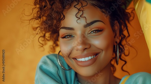 Joyful young woman with curly hair and hoop earrings smiling against a vibrant orange background