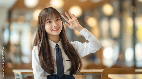 Asian girl in a sharp white Shirt sitting at her school desk and putting her index finger to her cheek and winking in a modernized classroom, blurred bokeh background