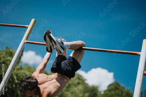 A young man working out in nature, performing calisthenics on a pull-up bar, showcasing physical strength and wellness under a clear blue sky.