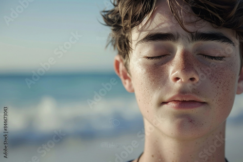 Close up of a tranquil young man meditating on the ocean shore, eyes closed, waves softly lapping in the background.