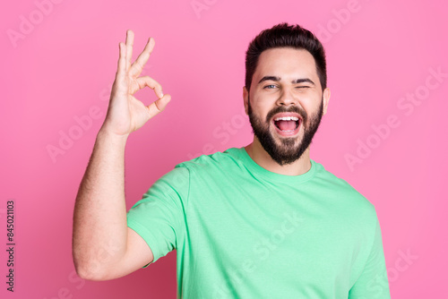 Photo of positive cheerful nice guy with stylish beard dressed green t-shirt showing okey winking eye isolated on pink color background