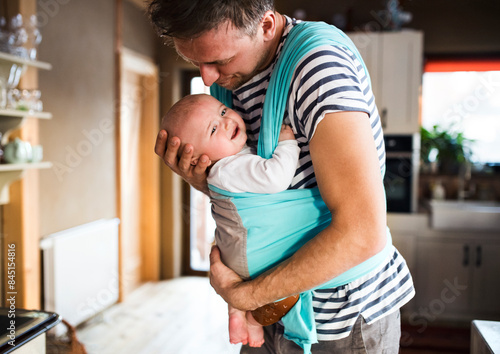 Father holding small baby, carrying him around house in baby carrier. Unconditional paternal love, father's Day.