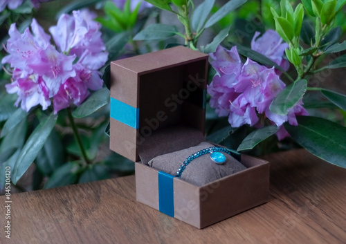 Niebieska nowoczesna i elegancka bransoletka z kryształów i ręcznie malowanej zawieszki ze szklanym kaboszonem w pudełku na biżuterię z liśćmi i różowymi kwiatami w tle