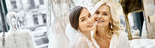 A young brunette bride in a wedding dress and her middle-aged blonde mother standing side by side in a bridal salon.