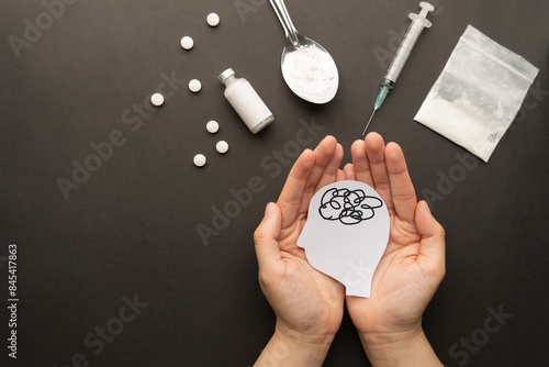 Drug addiction induced psychosis. Hands holding human brain feeling confused symbol with drug powder, syringe and pills on dark background. International Day against Drug Abuse.