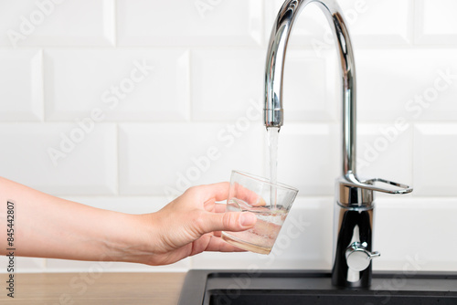 Thirst human wanted to drink water and filling up a glass of tap pure water from kitchen faucet with white tiles wall. Concept of purity and cleanness of fresh natural water with copy space