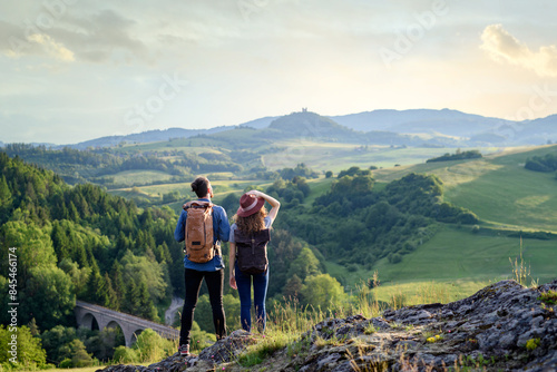 Travellers couple hiking on easy trail in nature with backpacks. Young tourist resting, enjoying breathtaking view. Summer vacation oudoors. Rear view.