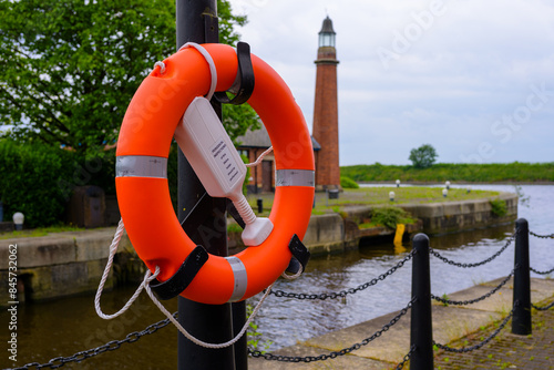 Orange lifebuoy on post by a river, brick lighthouse in the background on a cloudy day