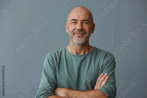 A confident, bald mature man wearing a green casual t-shirt smiles warmly with arms crossed against a monochromatic background