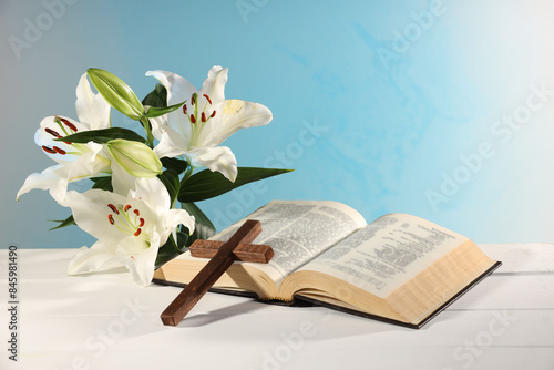 Wooden cross, Bible and beautiful lily flowers on white table against light blue background. Religion of Christianity