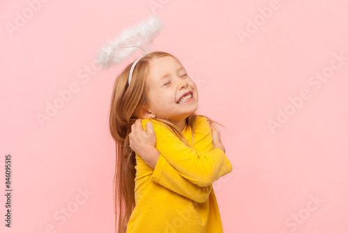 Portrait of cute smiling happy blonde little girl with nimb over head hugging herself with pleasure, self loving, wearing yellow jumper. Indoor studio shot isolated on pink background.