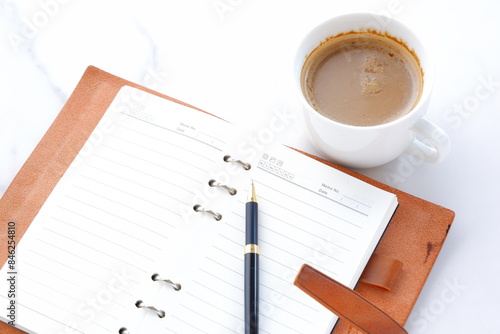 ミルクをたっぷり入れたコーヒーを飲みながら、茶色の革のシステム手帳を開いてデスクワーク中のイメージ 