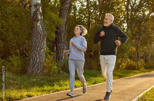 Happy smiling sporty senior couple wearing sportswear jogging in the park having sport workout. Elderly man and woman running outdoors. Workout jog in nature, exercising for retired concept.