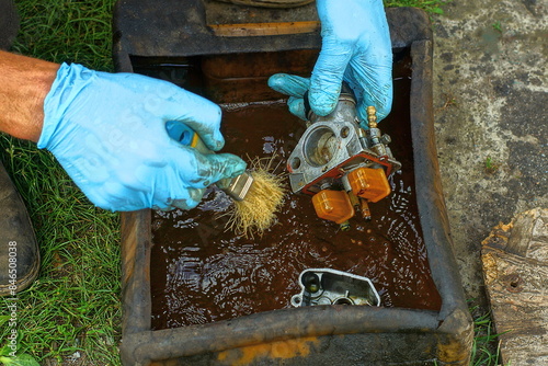 cleaning and washing with a brush in a plastic canister of gasoline with hands in blue rubber gloves a disassembled old dirty iron carburetor from a retro motorcycle during the day on the street