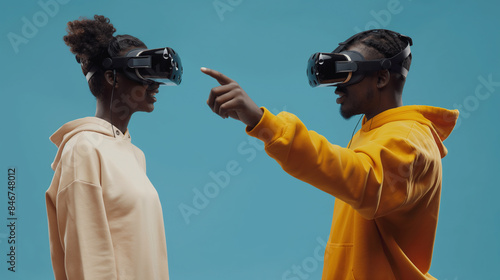 Homem e mulher africanos em moletons apontam um para o outro enquanto usam fones de ouvido VR em um fundo azul, foto de estoque para uma campanha publicitária
